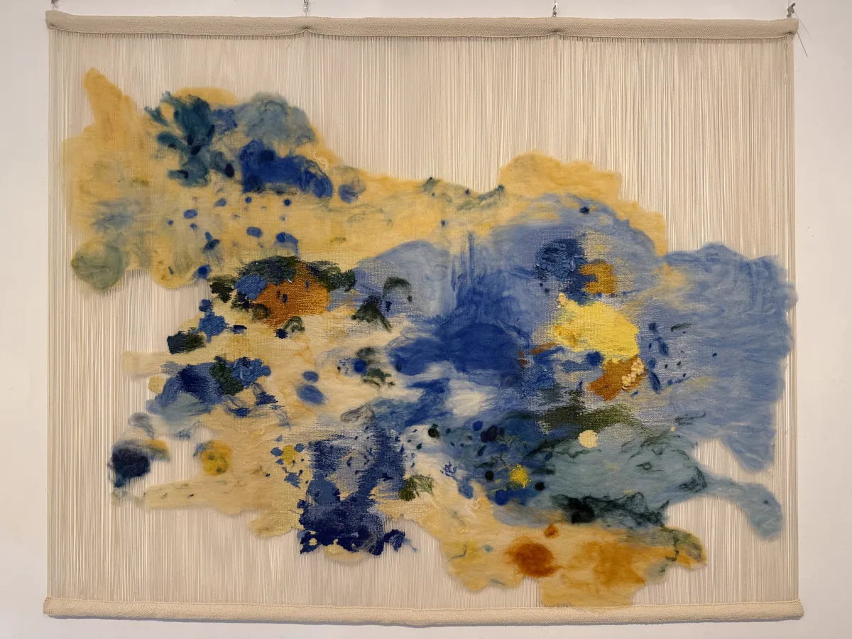 Large textile hanging: clouds of felted wool in cobalt blue, ochre and saffron drift across a field of bare cream warp threads stretched between two wooden bars.