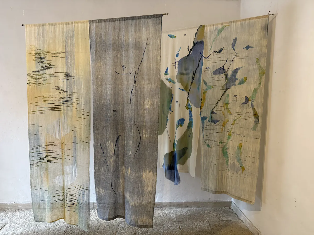 Four tall, translucent woven panels hang side by side in a whitewashed room, marked with ink-black stitched lines and washes of blue, green and yellow.