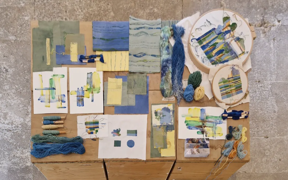 A studio table seen from above: woven and dyed samples, watercolour studies, skeins of blue and green yarn and two embroidery hoops.