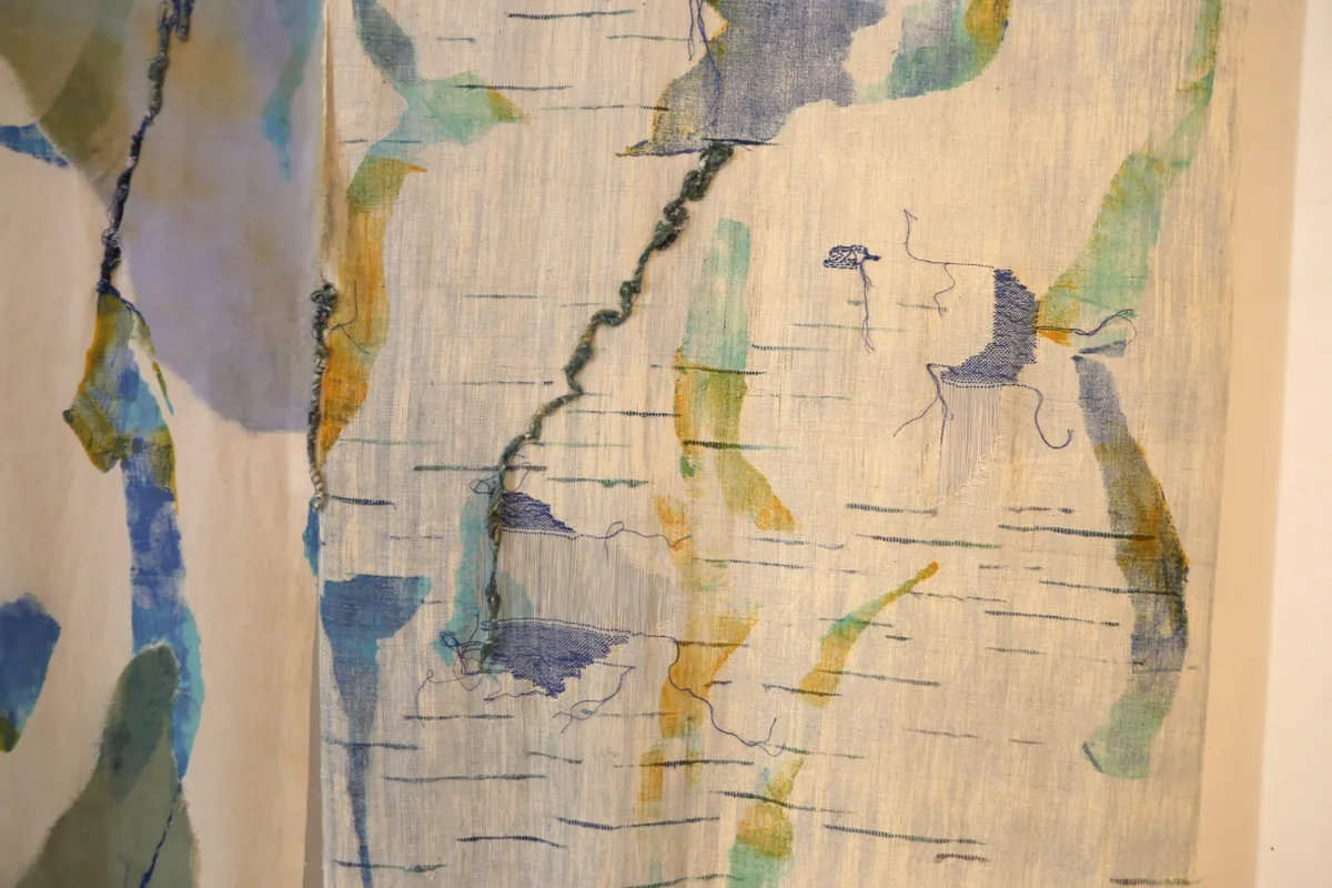 Detail of a sheer woven panel mended with blue and green stitches, a twisted yarn running across it.